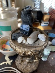 a blue-eyed raven totem carved out of obsidian sits on a fox-shaped pedestal with a piece of black tourmaline, a hagstone (stone with a naturally occurring hole in it), a piece of howlite (tumbled white stone with gray veins), and a tiny found bird skull, on a dusty desk cluttered with other tumbled stones, candles, and a small ceramic bowl containing strips of brightly colored watercolor paper.